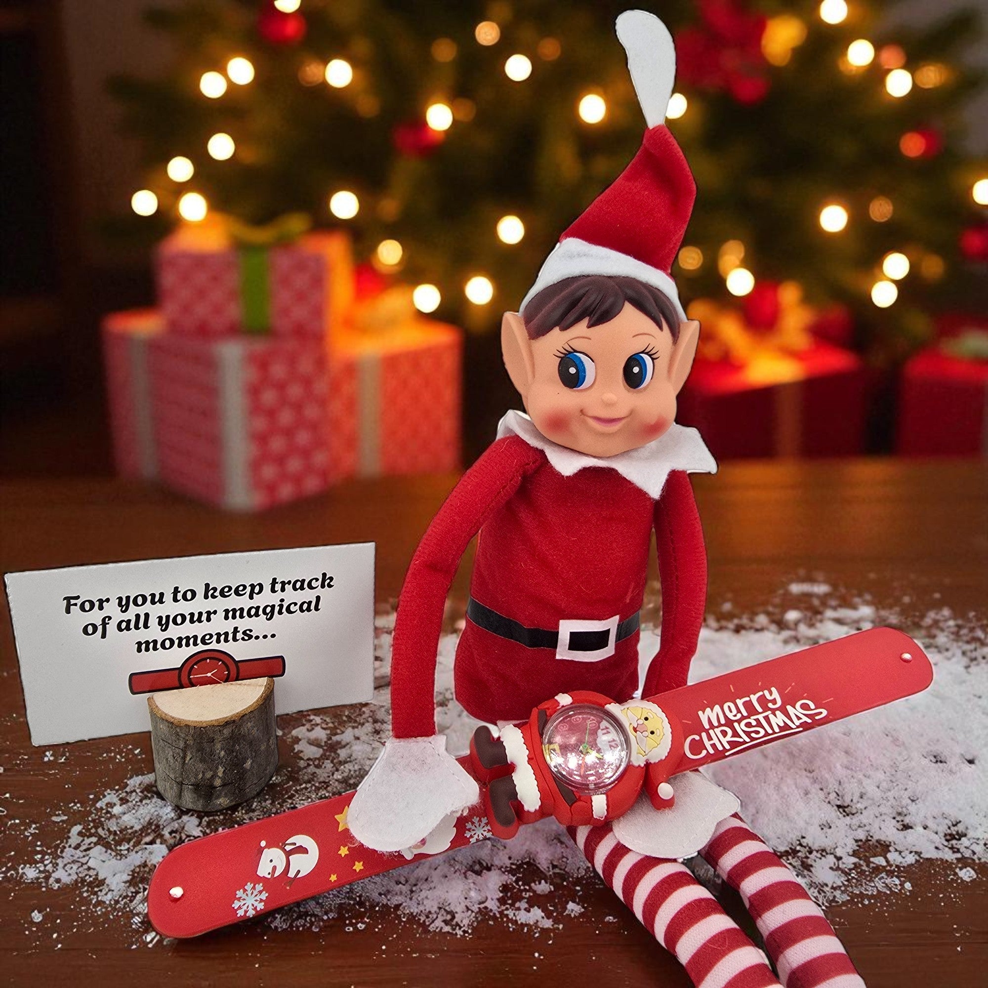 Christmas elf doll sitting in faux snow next to a red Merry Christmas slap wrist watch from the 24 day elf kit.
